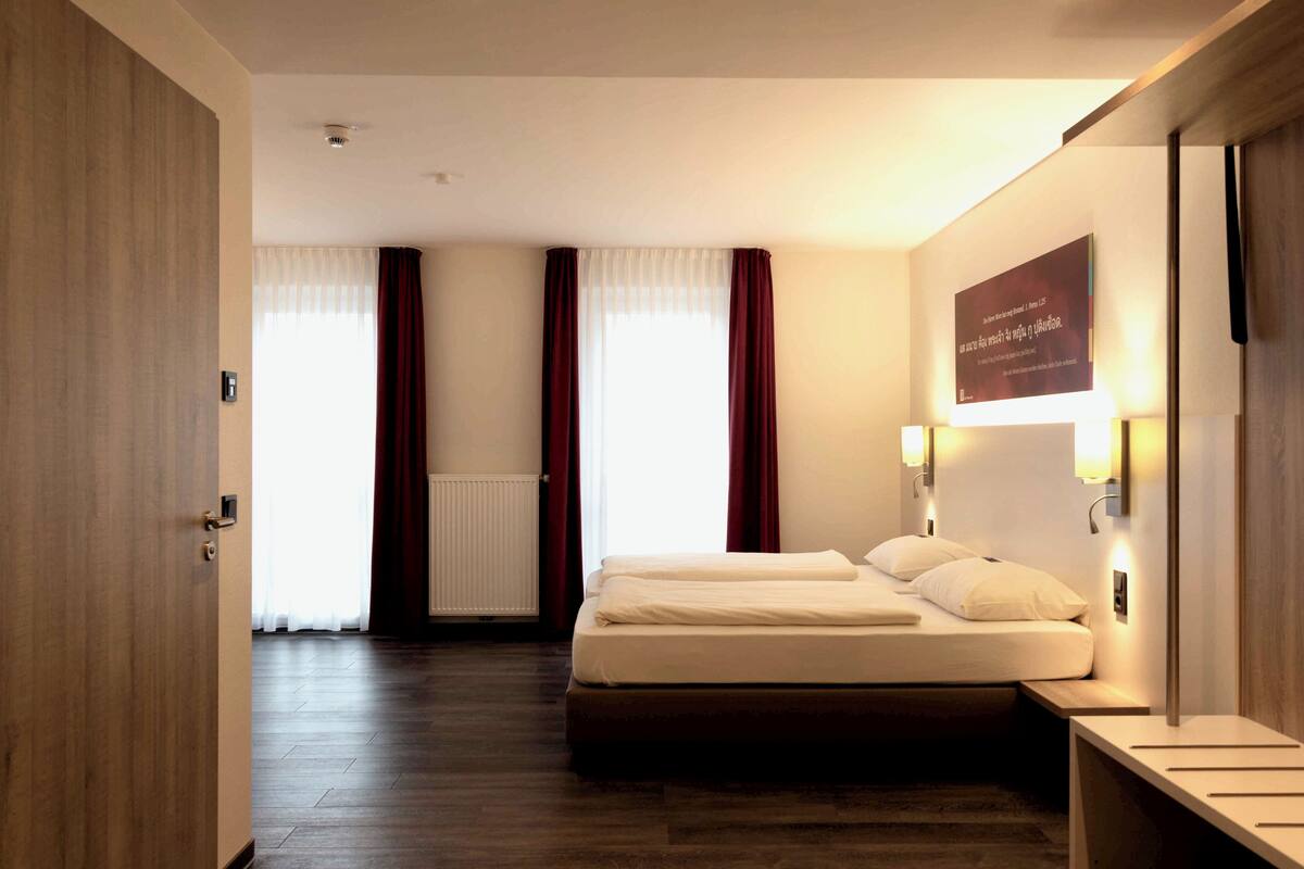 Ein Hotelzimmer mit Bett, Nachttisch, Wandlampe und einem Fenster mit Vorhängen.