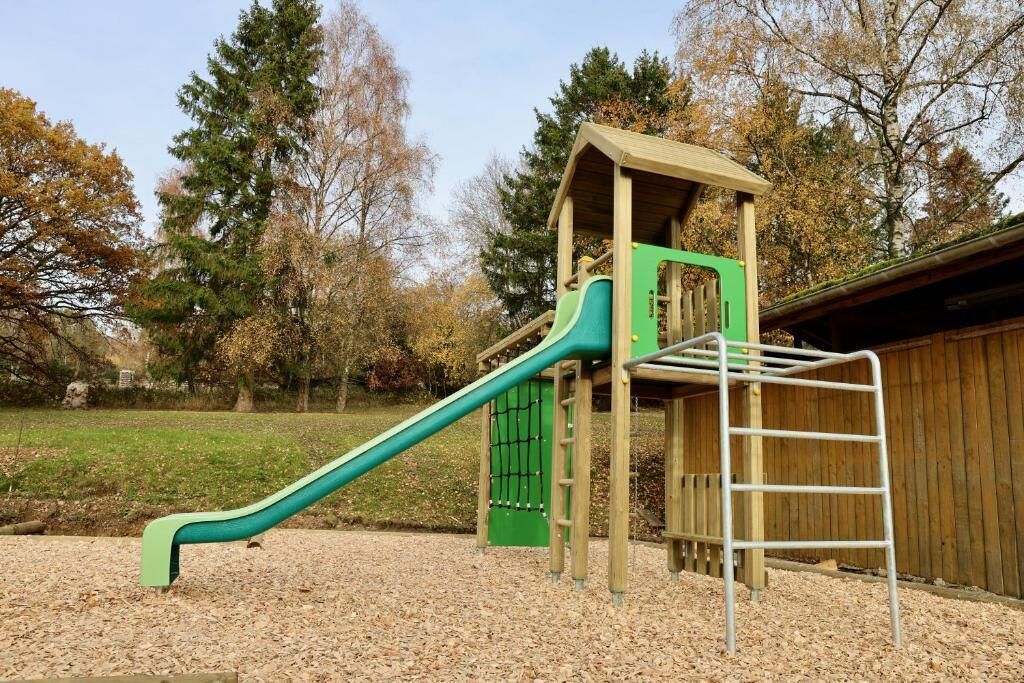 A playground with a slide and climbing area, surrounded by trees and a wooden fence.