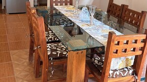Een eethoek met een glazen tafel, houten stoelen, een keuken op de achtergrond en een plafondventilator.