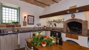 A kitchen with a brick oven, a wooden table with fresh produce, and a window with a view. - La Scorticata. Elegant family house with swimming pool, surrounded by oak trees. (Cura di Vetralla)