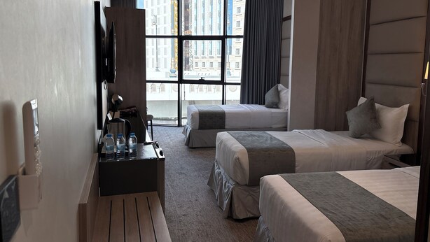 A hotel room with two beds, a desk with a chair, a TV, and a large window offering a city view. - SUQYA TAIBA (Madinah)