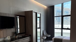 A modern hotel room with a large bed, a desk with a flat-screen TV, a chair, and a large window offering a city view. - SUQYA TAIBA (Madinah)