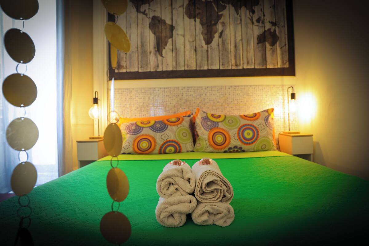 A neatly made bed with a green bedspread, two rolled towels, and patterned pillows.