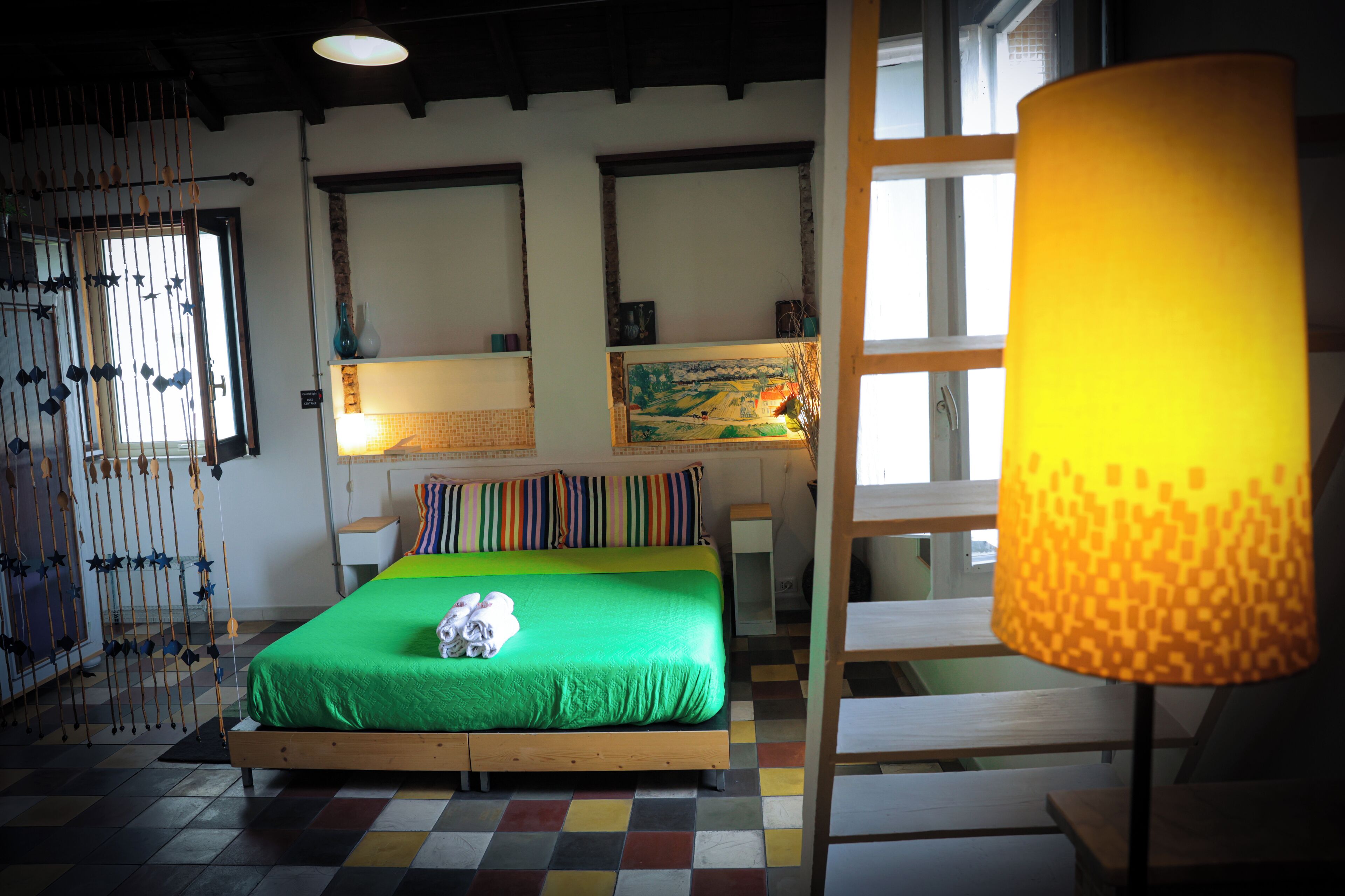 A room with a green bed, a wooden floor, a yellow lamp, and a window with a view.