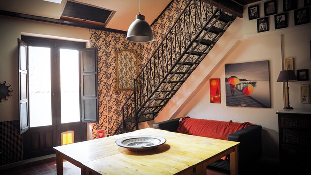 Ein Zimmer mit einem Holztisch, einem schwarzen Sofa, einer Treppe mit schmiedeeisernem Geländer und einem Bild an der Wand.