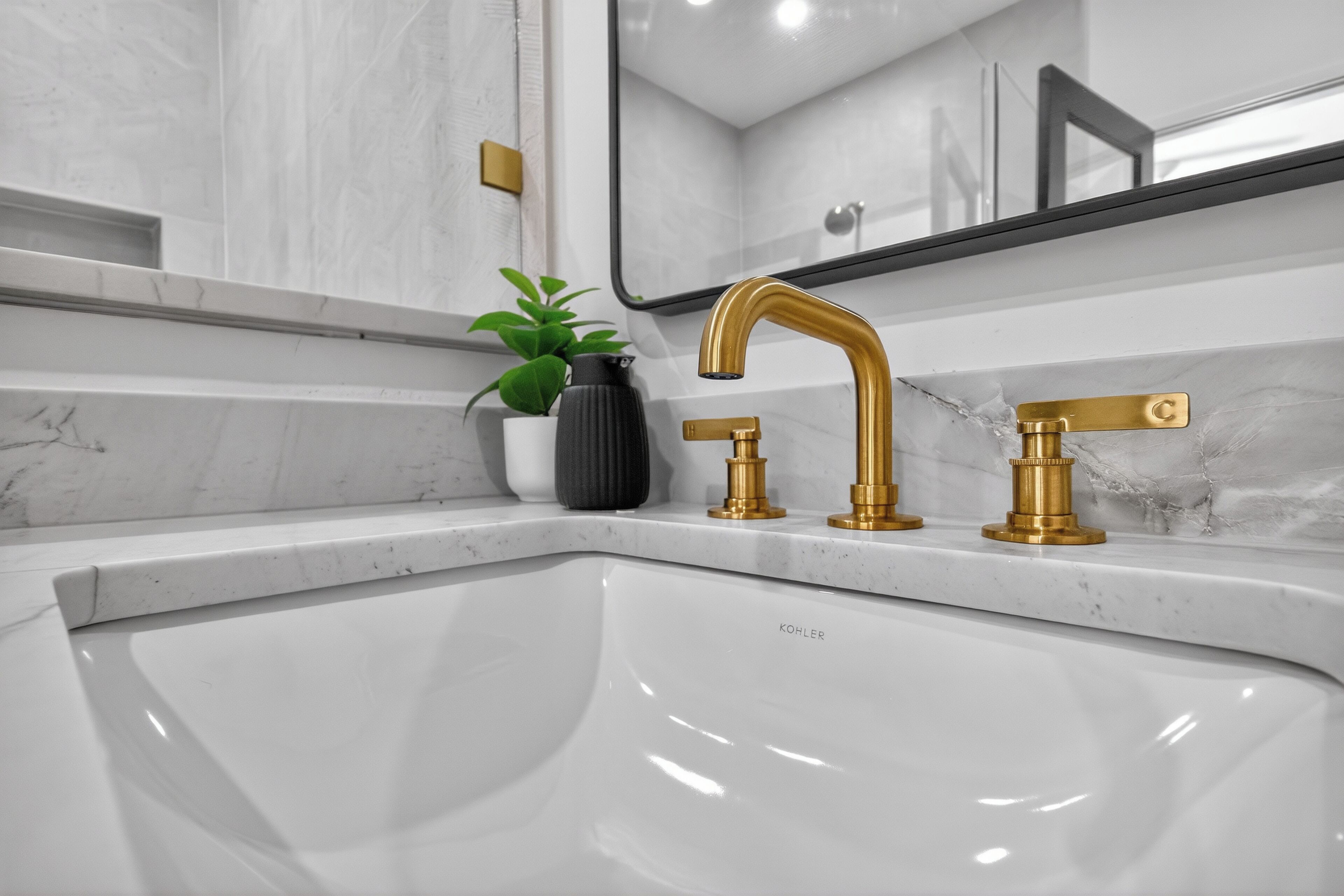 Ein modernes Badezimmer mit einem weißen Waschbecken, goldenen Wasserhähnen und einer schwarzen Vase mit einer Pflanze.