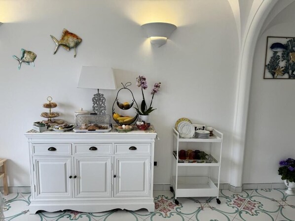 Un meuble blanc avec des tiroirs, un panier suspendu, une lampe et une petite étagère avec de la vaisselle.