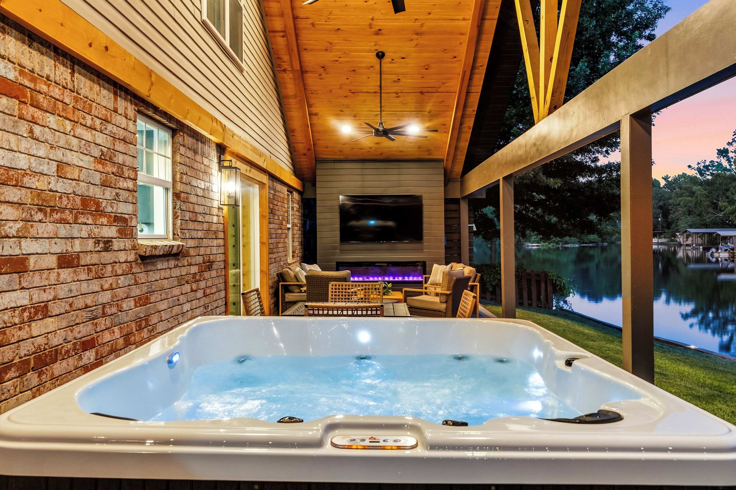 A hot tub with a flat-screen TV and outdoor seating, surrounded by a wooden deck and a brick wall.