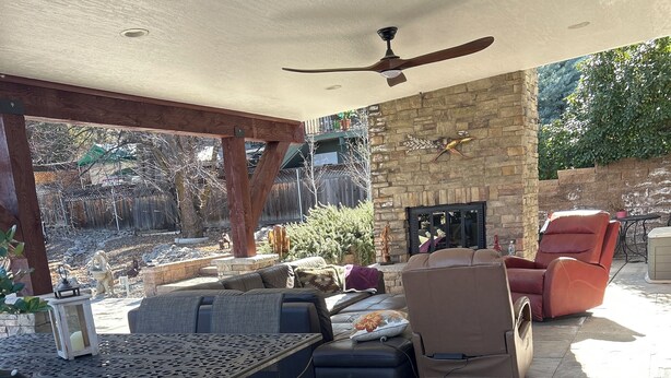 Un patio al aire libre con chimenea de piedra, ventilador de techo y diversas disposiciones de asientos, incluyendo un sofá, sillas y una mesa.