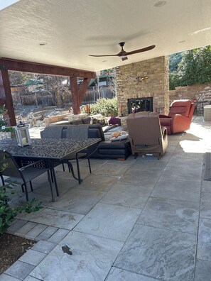 Une terrasse extérieure dotée d’une cheminée en pierre, d’un ventilateur de plafond et de différents types de mobilier, notamment un canapé, des fauteuils et une table.