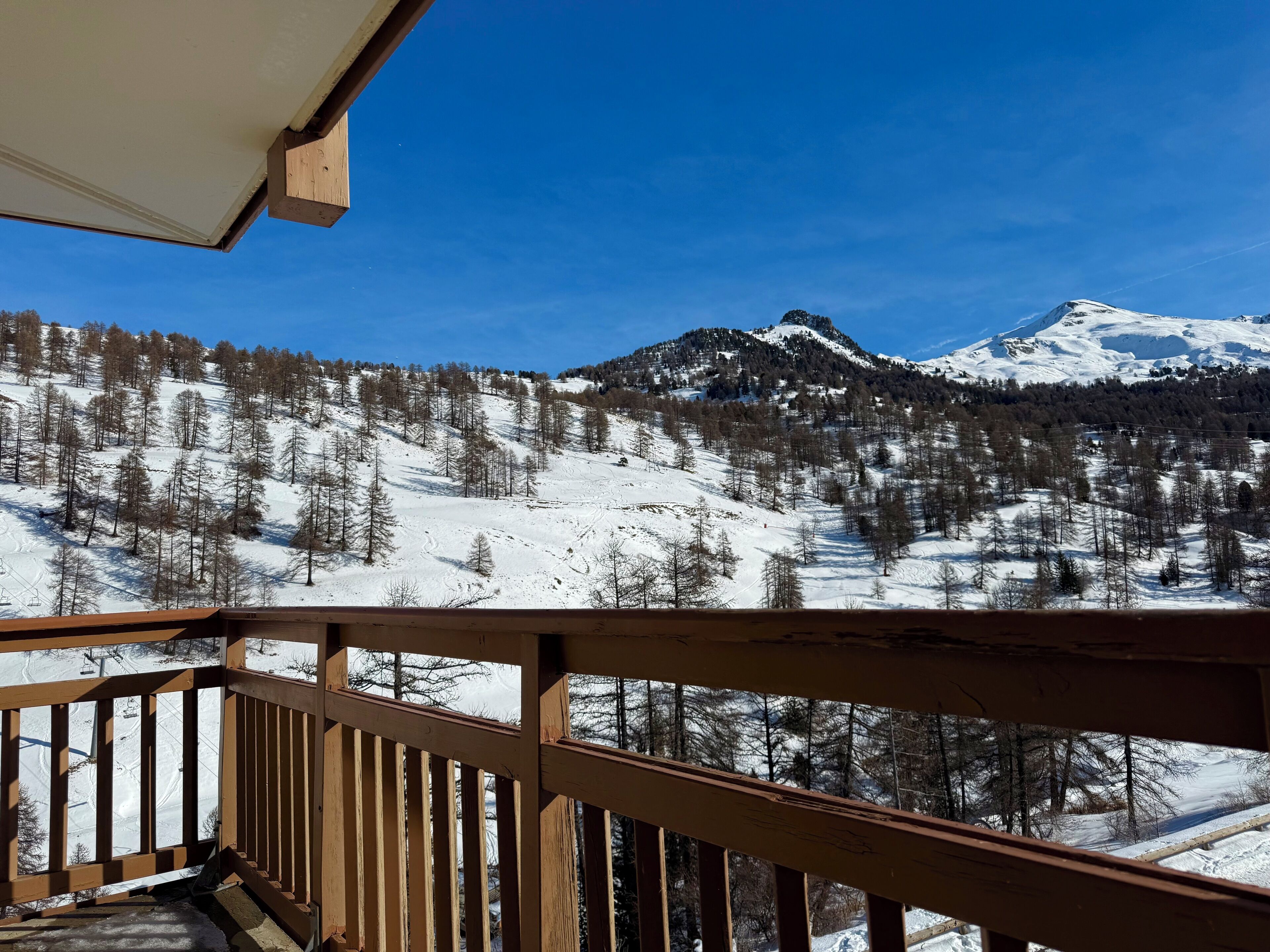 A wooden balcony with a view of a snow-covered mountain range and a clear blue sky.