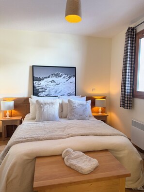 A neatly made bed with fluffy pillows and a blanket, a wooden bedside table with a towel, a framed picture of a snowy mountain on the wall, and two bedside lamps. - Luxury half chalet 95 m2, ski-in/ski-out, panoramic view, indoor pool (Vars)
