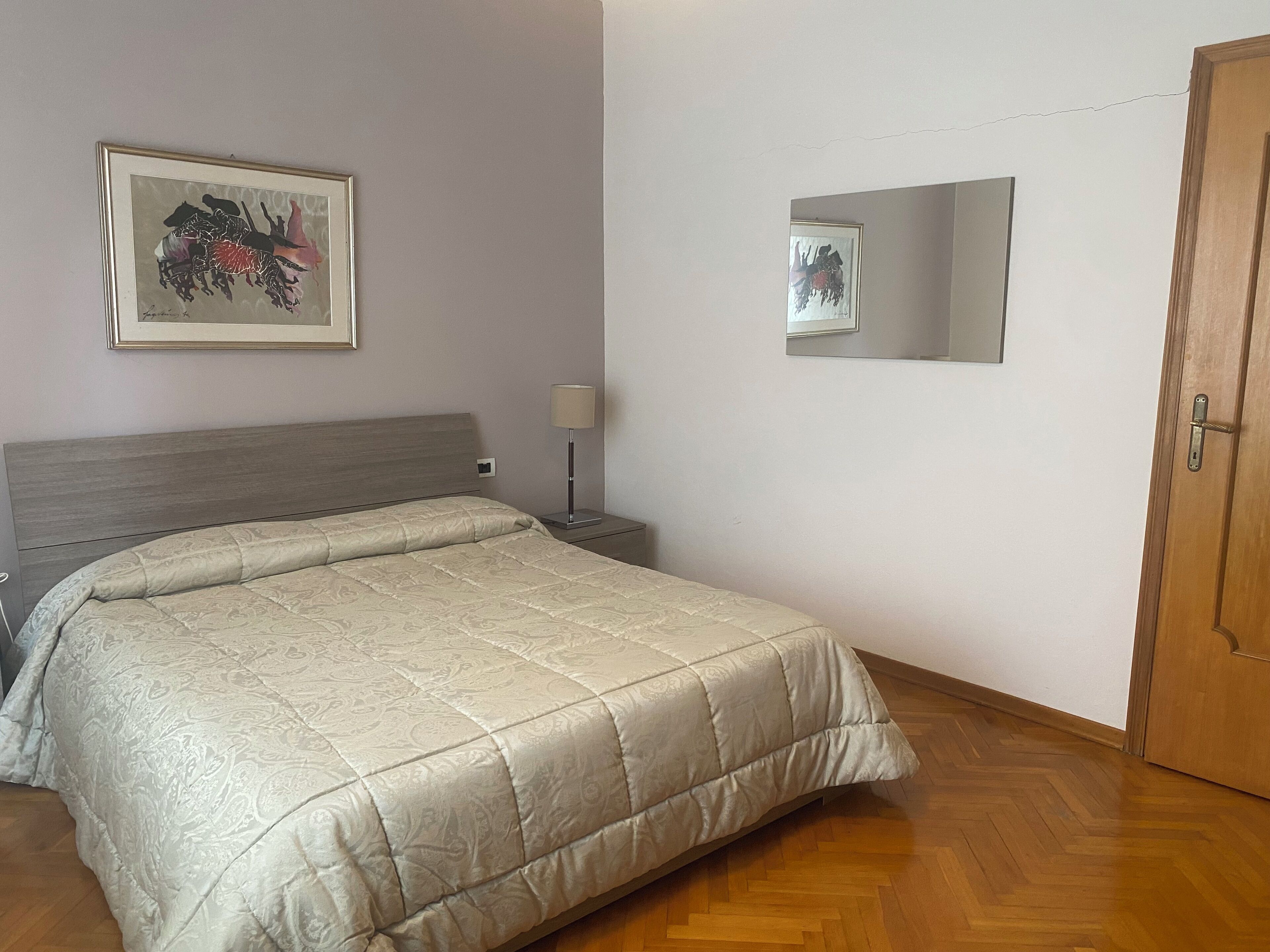 Een netjes opgemaakt bed met een bedspreider met een patroon, een bedlamp en een ingelijst foto aan de muur.