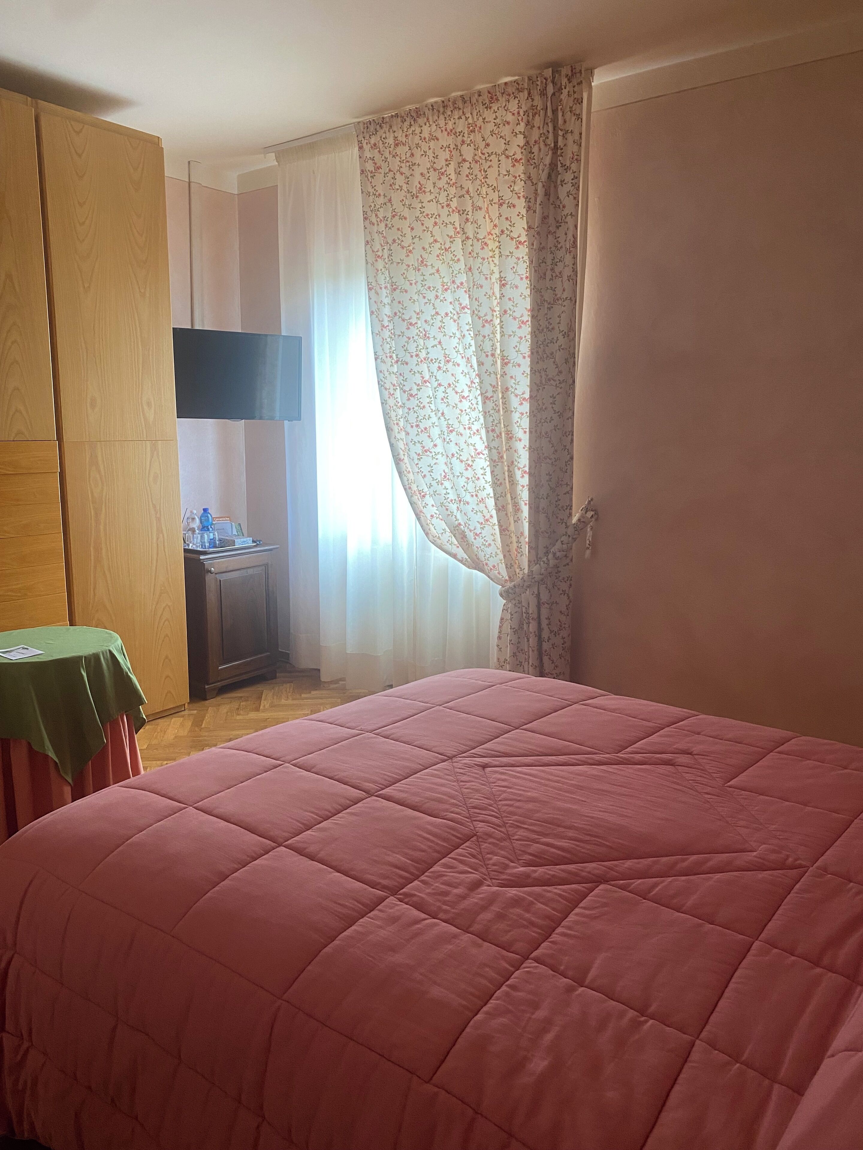 Un dormitorio con una cama grande, un armario de madera, una mesita con mantel verde y una kitchenette al fondo.