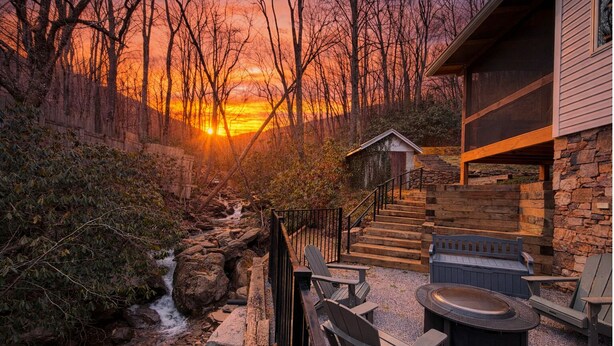 A stone staircase leading to a wooden deck with outdoor seating, overlooking a stream and a sunset. - New! Private Waterfall! King Suite~Pet Friendly (Burnsville)