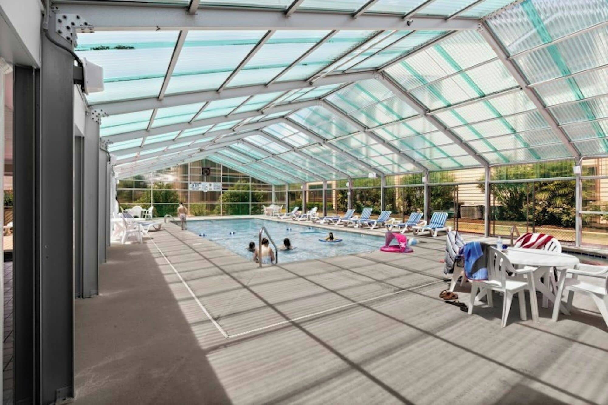 A covered swimming pool with clear glass panels, lounge chairs, and a poolside area.