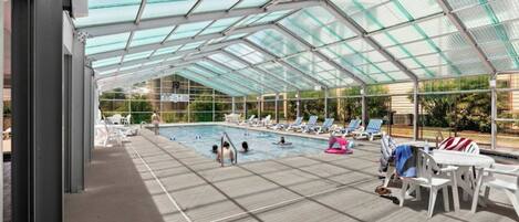 A covered swimming pool with clear glass panels, lounge chairs, and a poolside area.