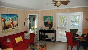A living room with a red sofa, yellow armchair, and a glass coffee table. There is a dining area with a table and chairs, and a ceiling fan. - Guest Cottage 20- 2BR House- Community Pool (Myrtle Beach)