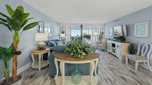 Een ruime woonkamer met een grote plant, een ronde houten tafel en een bank. Er is een zithoek met een tafel en stoelen, en een keuken met een kast en een stoel.