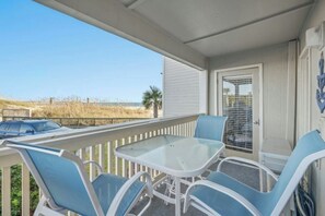 Un balcon avec une table en verre et des chaises bleues, donnant sur une plage et une route.
