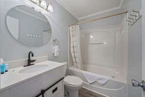 Une salle de bain avec un lavabo blanc, un miroir et des toilettes. On y trouve un rideau de douche et un porte-serviettes.