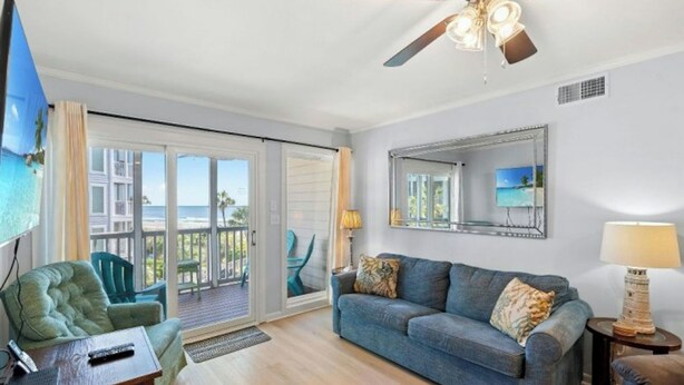 A living room with a blue sofa, a green armchair, a ceiling fan, and a large window overlooking a balcony with a view of the ocean. - Ocean View 3BR At Pelican's Watch 209- Pool (Myrtle Beach)