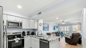 A modern kitchen with white cabinets, a microwave, an oven, and a dishwasher. There is a dining area with a table and chairs, and a living room with a couch and a TV. - Ocean View 3BR At Pelican's Watch 209- Pool (Myrtle Beach)