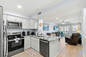 A modern kitchen with white cabinets, a microwave, an oven, and a dishwasher. There is a dining area with a table and chairs, and a living room with a couch and a TV. - Ocean View 3BR At Pelican's Watch 209- Pool (Myrtle Beach)