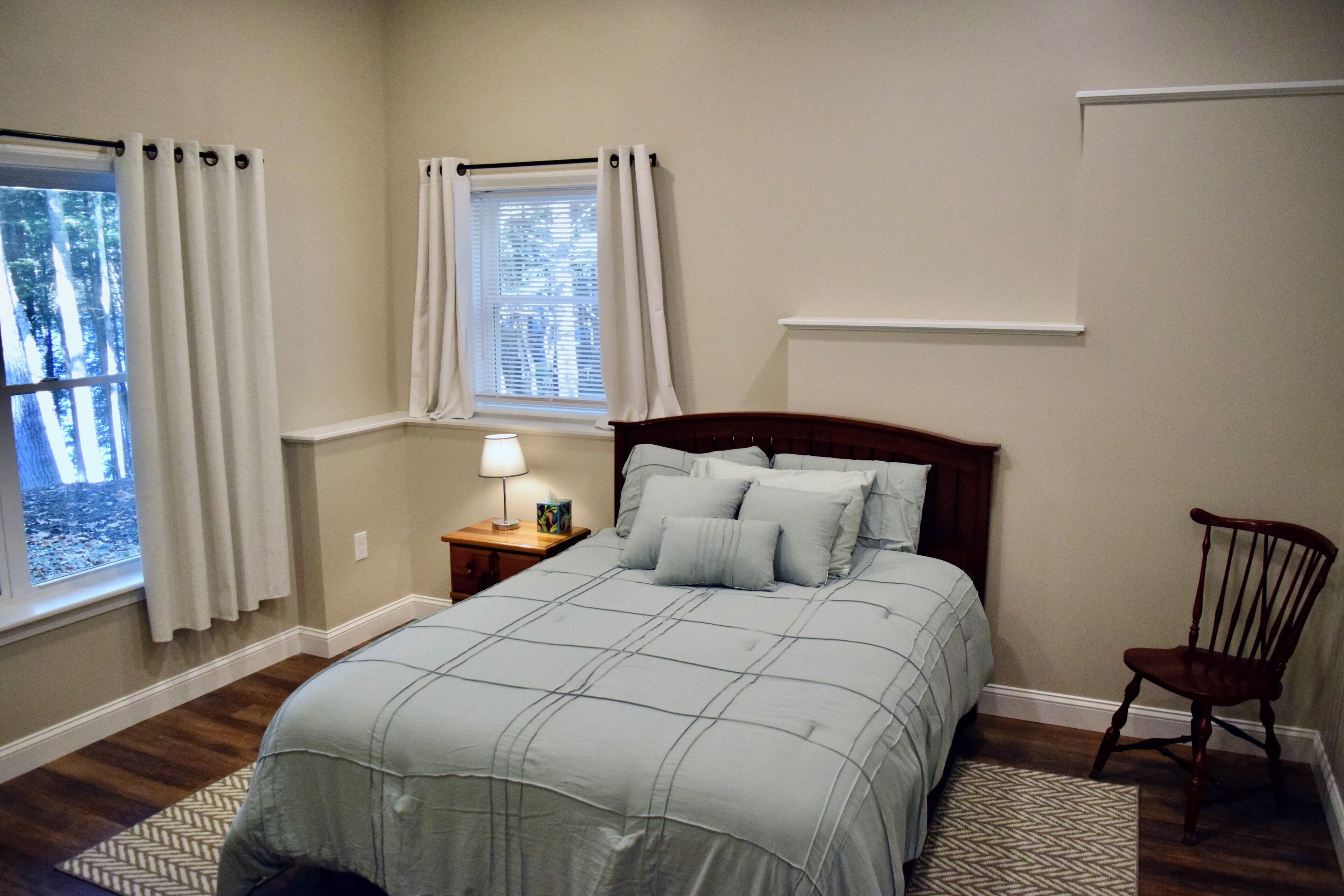 A bedroom with a bed, nightstand, chair, and two windows with curtains.