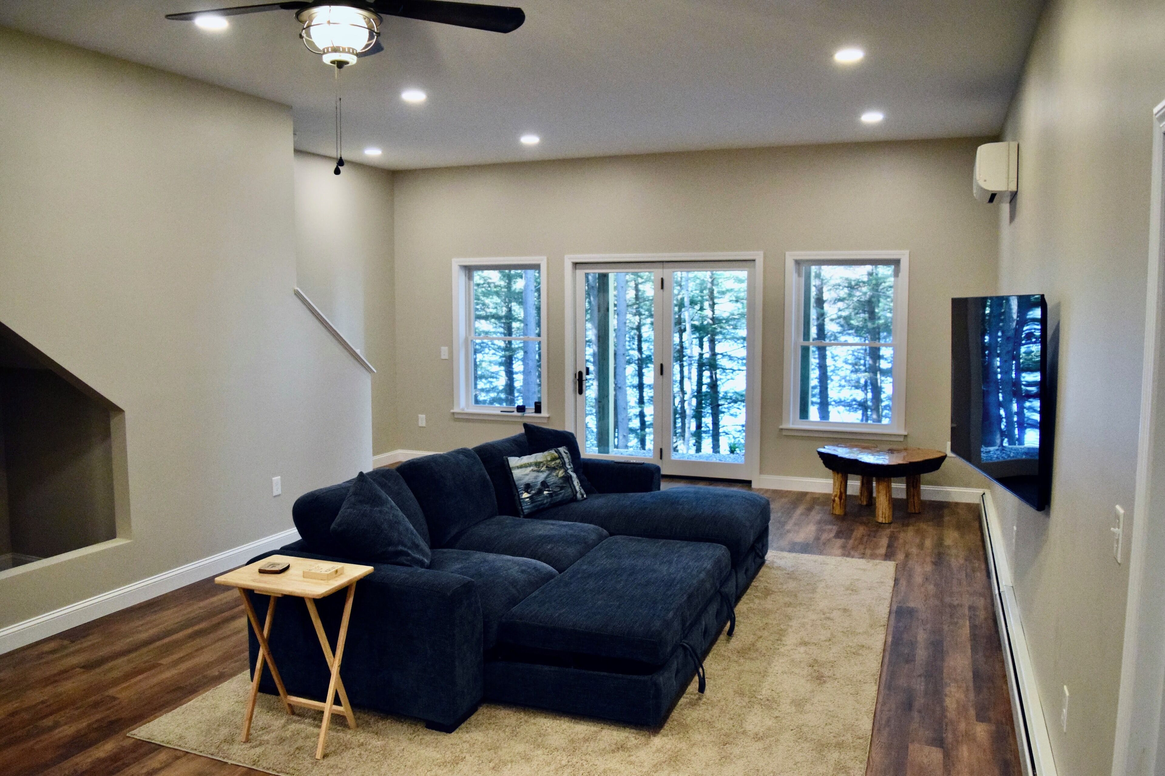 A living room with a sectional sofa, a small wooden table, and a fireplace. There are large windows with a view of trees.