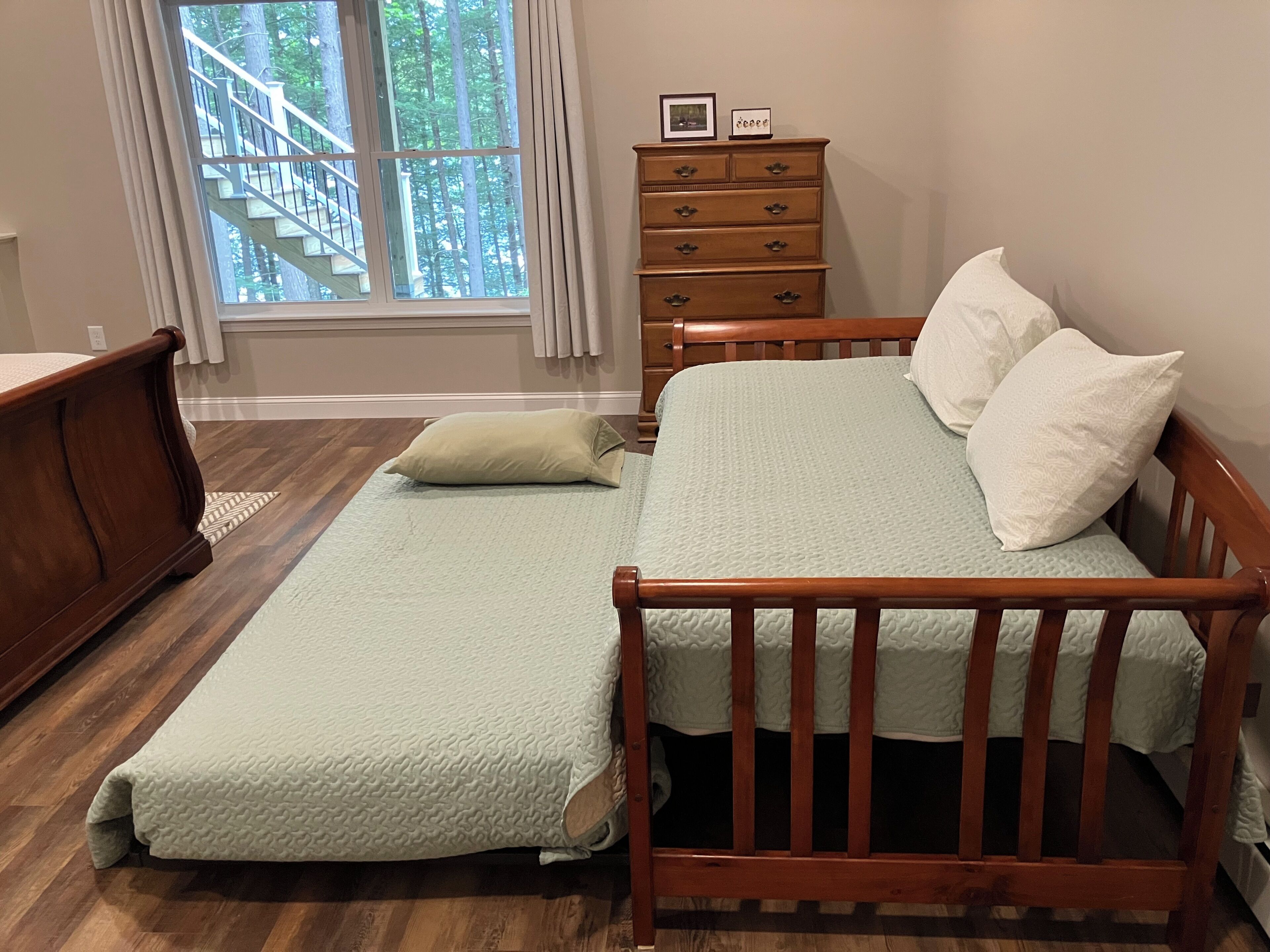 A bedroom with a wooden bed, a wooden dresser, a window with a view of a staircase and trees, and a light-colored floor.