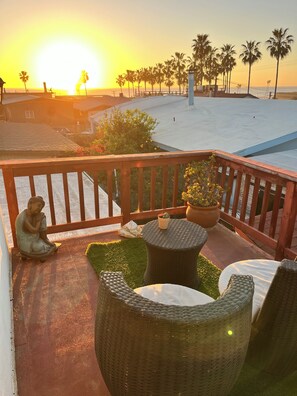 Un balcon agrémenté de meubles en osier, d’une statue de Bouddha et offrant une vue sur la plage et les palmiers au coucher du soleil.