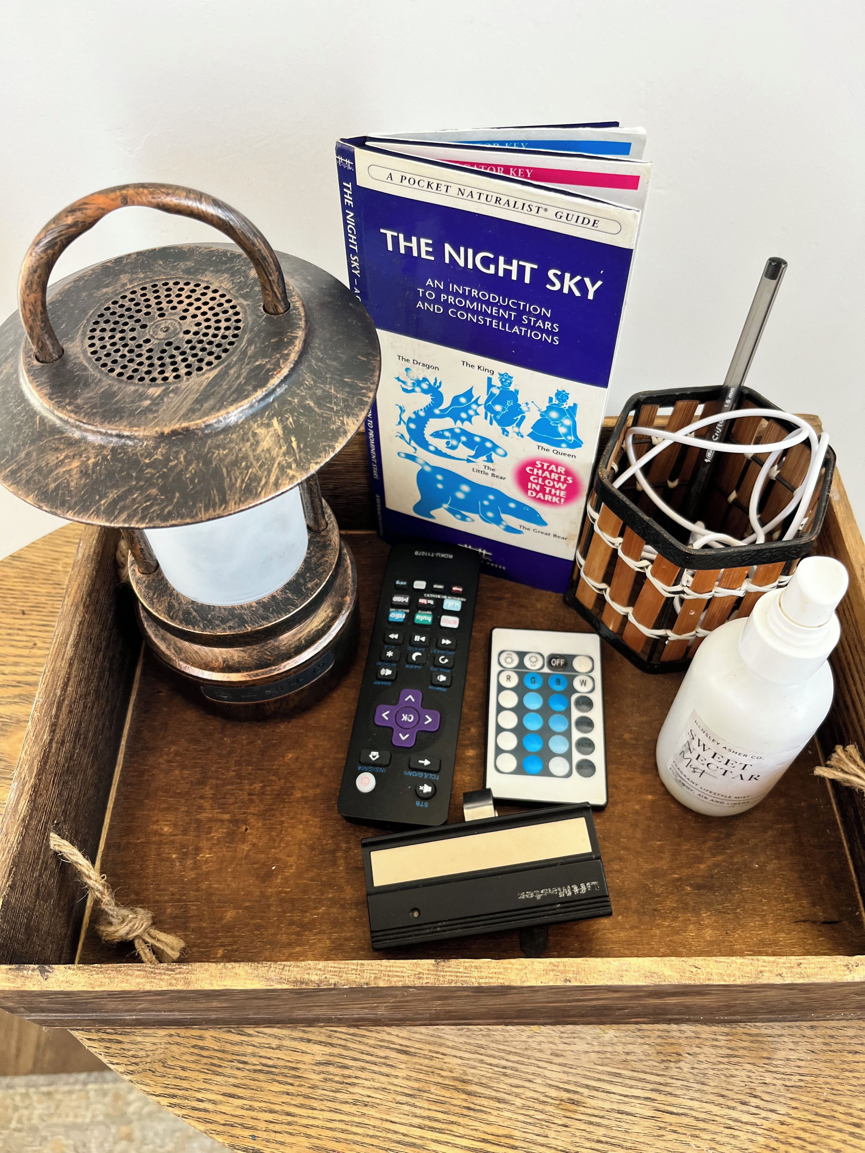 Ett träbricka med en lampa i vintage-stil, en fjärrkontroll, en bok med titeln ”Nattens himmel”, en dekorativ artikel som liknar en korg och en flaska med doftande spray.