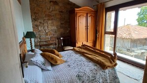 A bedroom with a stone wall, wooden ceiling beams, a bed with a patterned bedspread, a wooden wardrobe, and a window with a view of a building outside. - Nordés - Rural house in Lastres between the sea and the mountains (Luces)