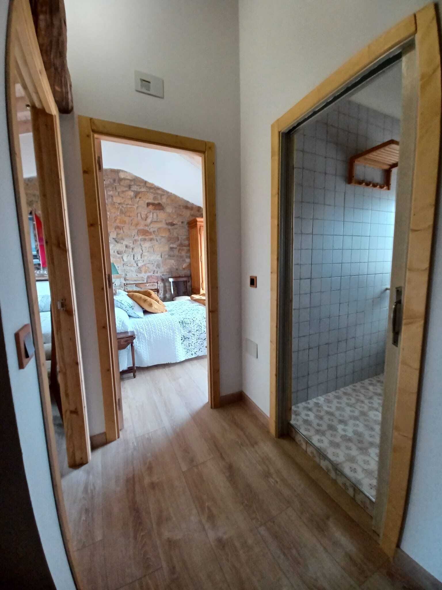 A narrow hallway with a wooden door leading to a bedroom with a stone wall and a bed with a white bedspread.