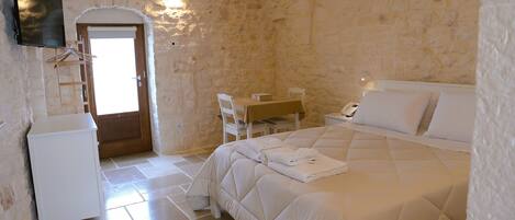 A bedroom with a stone wall, a bed with a quilt, a desk with a chair, and a television mounted on the wall.
