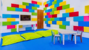 Een kamer met een blauw tapijt, een houten deur, een witte bank, een tafel met stoelen, en kleurrijke foam blokken aan de muren.