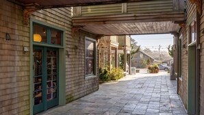 Une allée couverte, avec un revêtement en bois et un pavage en pierre, mène à un bâtiment doté d’une porte verte et de fenêtres.