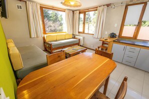 Una habitación compacta con una mesa de comedor de madera, un sofá, una pequeña cocina y una ventana con cortinas.