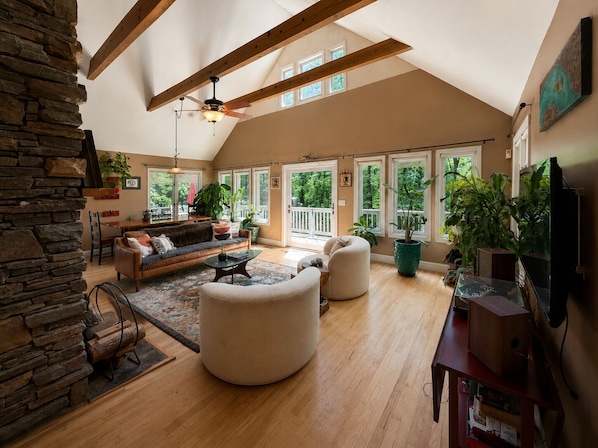 A spacious living room with a high ceiling, wooden beams, a stone wall, a sofa, a coffee table, and large windows.