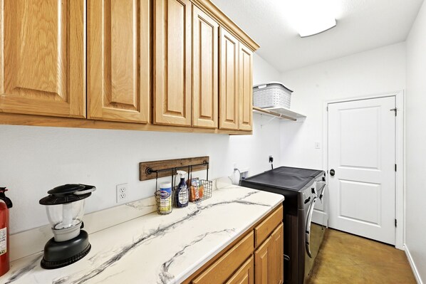 A kitchen with wooden cabinets, a marble countertop, a blender, a washing machine, and a fire extinguisher.