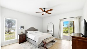 Quarto com cama, mesa de cabeceira, abajur, ventilador de teto e uma janela grande com vista para o exterior.