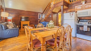 Interior acogedor de una cabaña con una mesa de comedor de madera, sillas de mimbre, un sofá, una chimenea, un televisor y una zona de cocina con refrigerador y estufa.