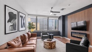 Un salon moderne avec un canapé en cuir, un pouf rond, un fauteuil noir, un téléviseur fixé sur un mur en bois et une vue sur la ville à travers de grandes fenêtres.