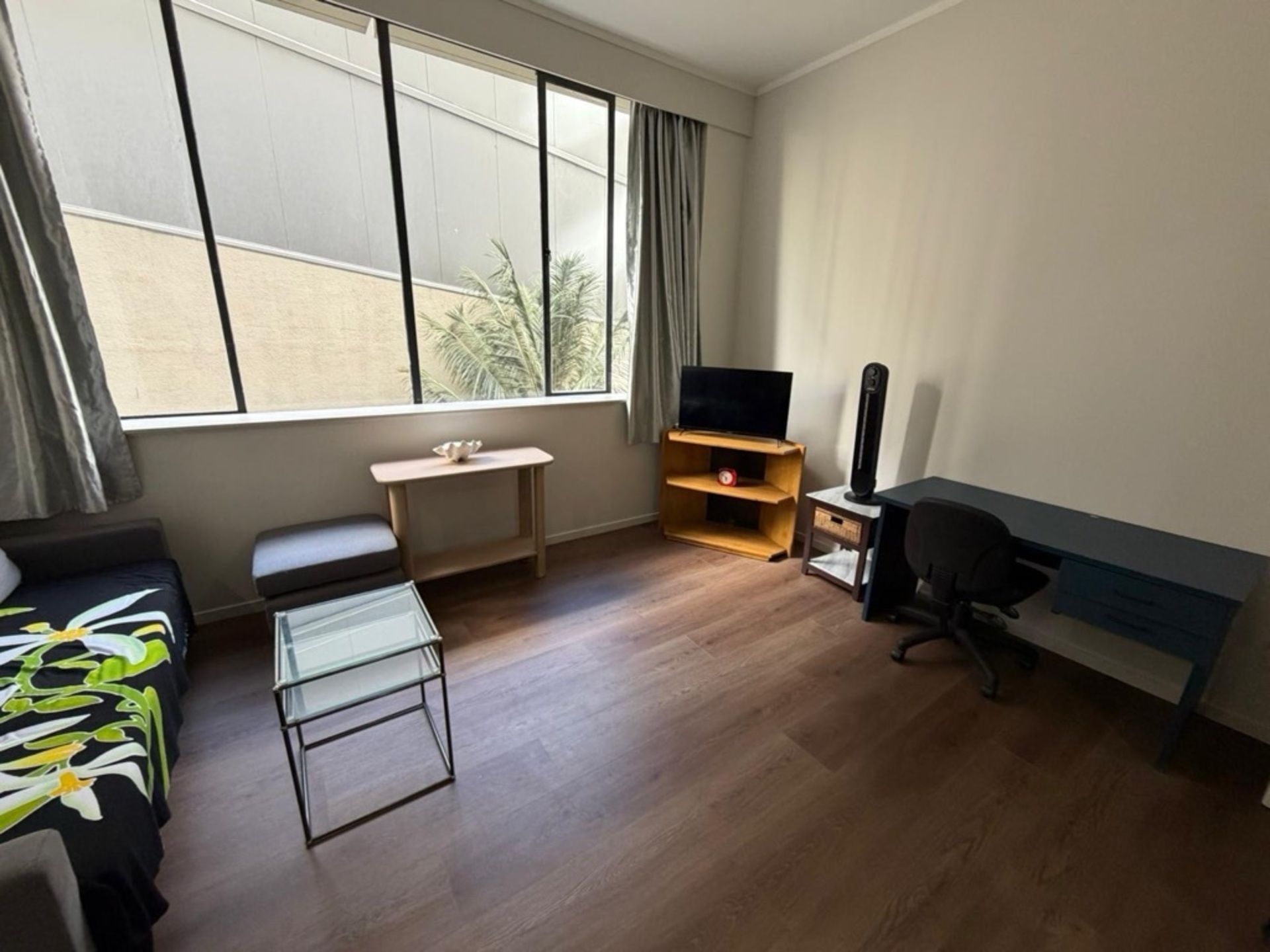 A modern hotel room with a sofa, a glass coffee table, a TV on a wooden stand, a desk, and a chair.