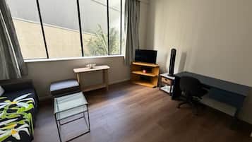 A modern hotel room with a sofa, a glass coffee table, a TV on a wooden stand, a desk, and a chair.