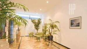 Un hall d’entrée spacieux avec un plafond haut, une grande plante en pot, un coin salon avec des fauteuils et un tableau encadré au mur.