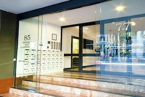 Entrada de un hotel con fachada de vidrio, carteles de apartamentos numerados y una escalera visible que conduce al interior.