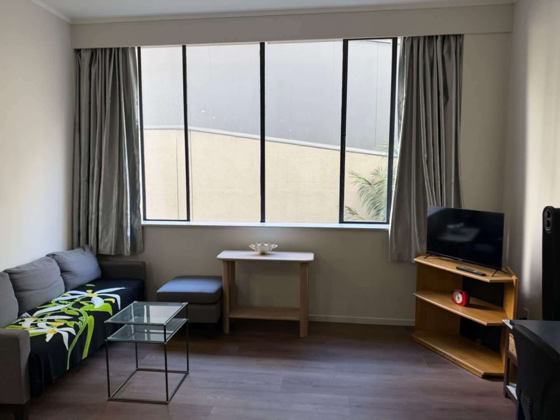 A living room with a sofa, a glass coffee table, a TV on a wooden stand, a small table with a white object, and a window with curtains.