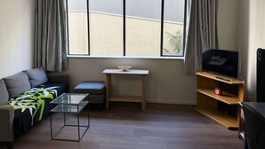 Un salon avec un canapé, une table basse en verre, un téléviseur posé sur un meuble en bois, une petite table sur laquelle se trouve un objet blanc, et une fenêtre avec des rideaux.